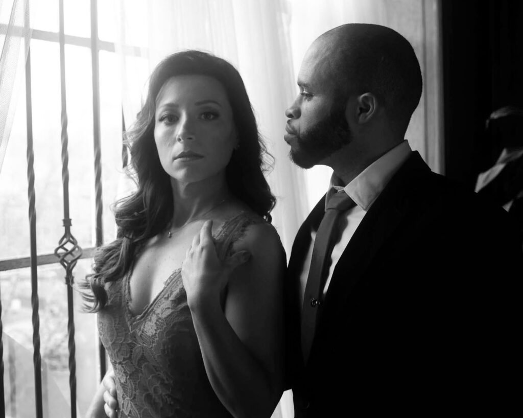man and woman window light black and white portrait photo she is wearing a dress he is wearing a suit and tie.