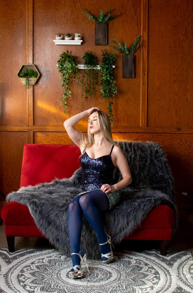 Blonde mandii monarch posing in studio. woman sexy in blue shimmer gown and stockings with heels holding hair in sunlight on couch.